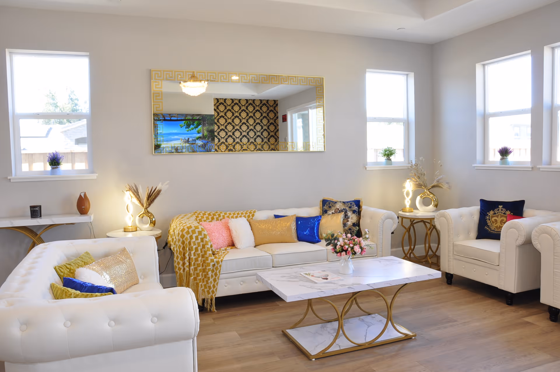 Bright furnished living room with white tufted sofas, colorful throw pillows, a marble-top coffee table and a large decorative mirror.
