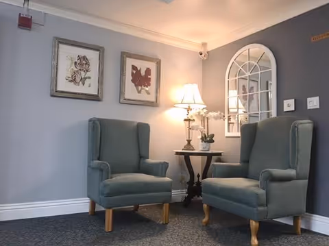 A cozy sitting area with two green upholstered armchairs facing each other, a small round wooden table between them holding a lamp and a white orchid plant. The walls are painted light gray with two framed floral artworks and a decorative window-style mirror hanging above the chairs.