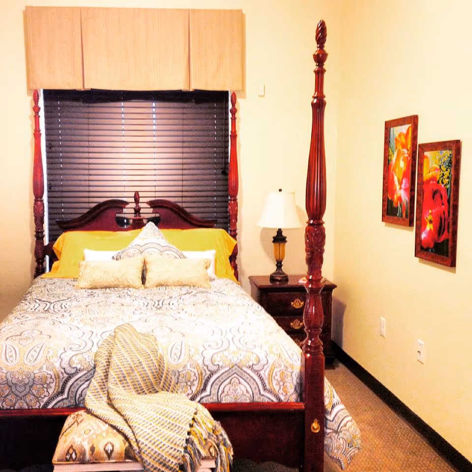 A furnished bedroom with a four-poster bed, patterned bedding, a bedside table and lamp, and floral wall art on the wall.