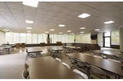 A spacious dining room with multiple rectangular tables and chairs arranged in rows. Large windows along the far wall allow natural light to fill the room. The ceiling has recessed lighting, and there is a door visible on the right side leading outside or to another area.