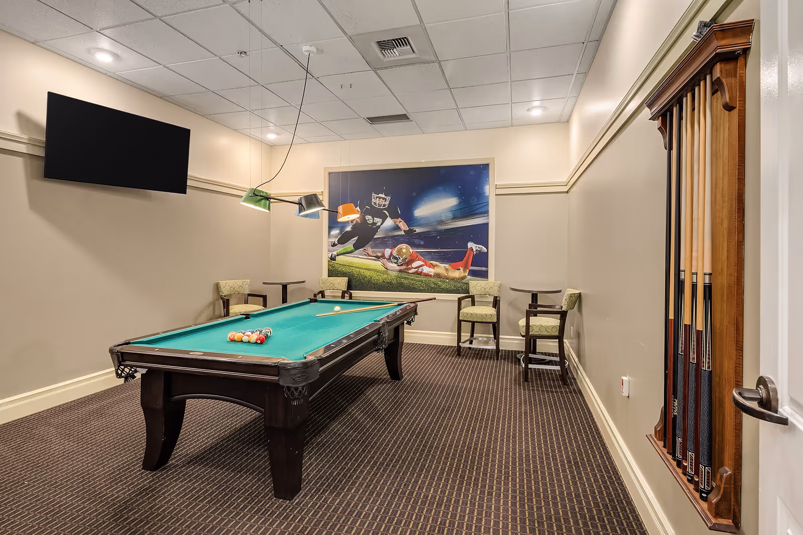A game room with a pool table in the center, a rack of pool cues mounted on the wall to the right, a flat-screen TV mounted on the left wall, and a large sports-themed mural featuring football players on the far wall. There are also two small tables with chairs along the back wall.