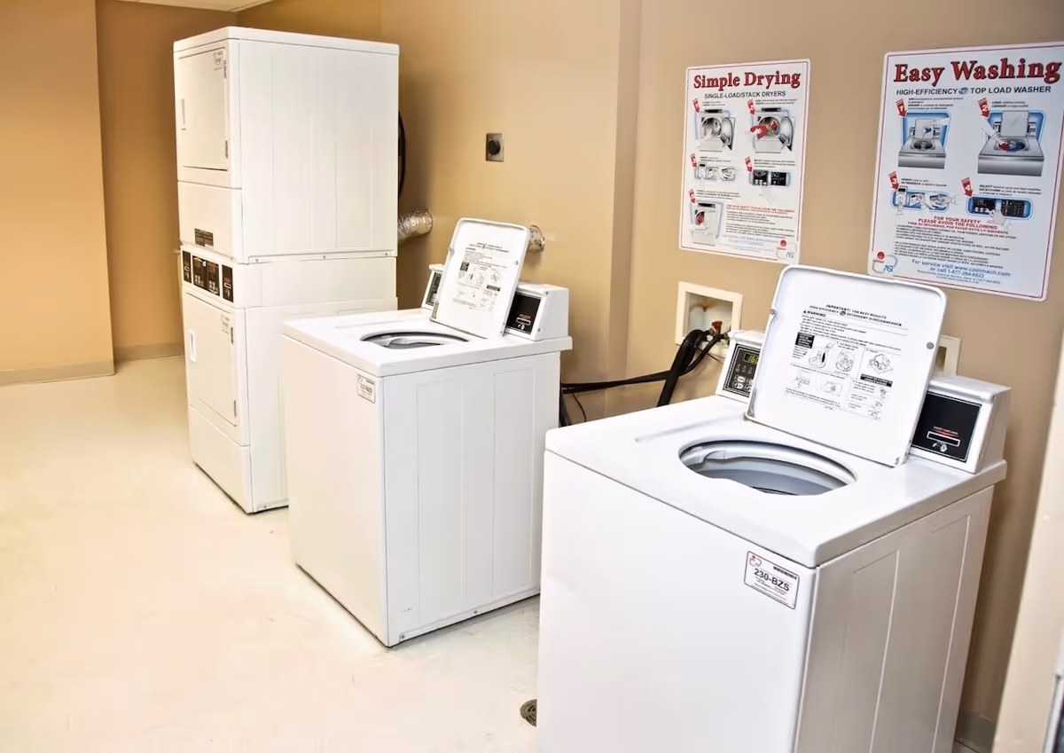 Laundry room with two top-loading washing machines and a stacked washer and dryer unit against beige walls. Instructional posters for drying and washing are displayed on the wall above the machines.