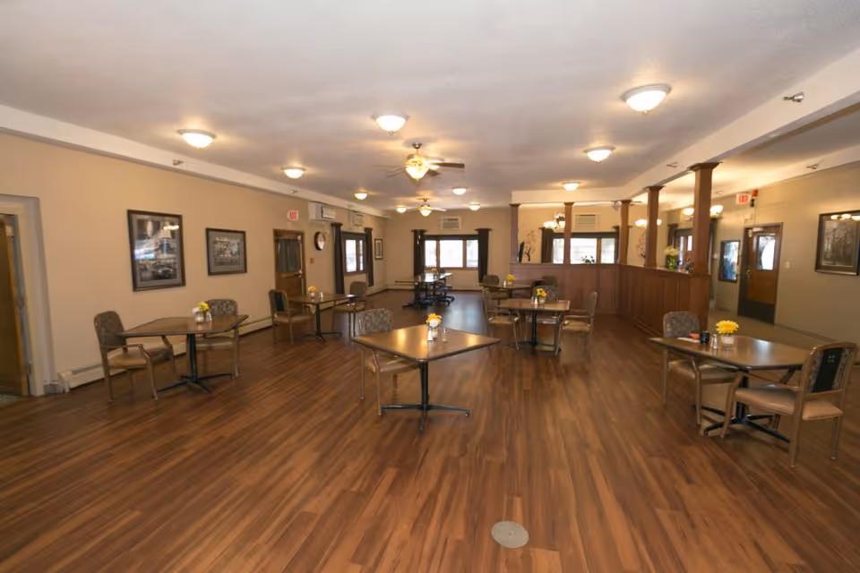 A spacious dining room with several square tables and chairs arranged neatly on a wooden floor. The room has beige walls adorned with framed pictures, multiple ceiling lights, ceiling fans, and windows letting in natural light. There are small flower arrangements on each table.