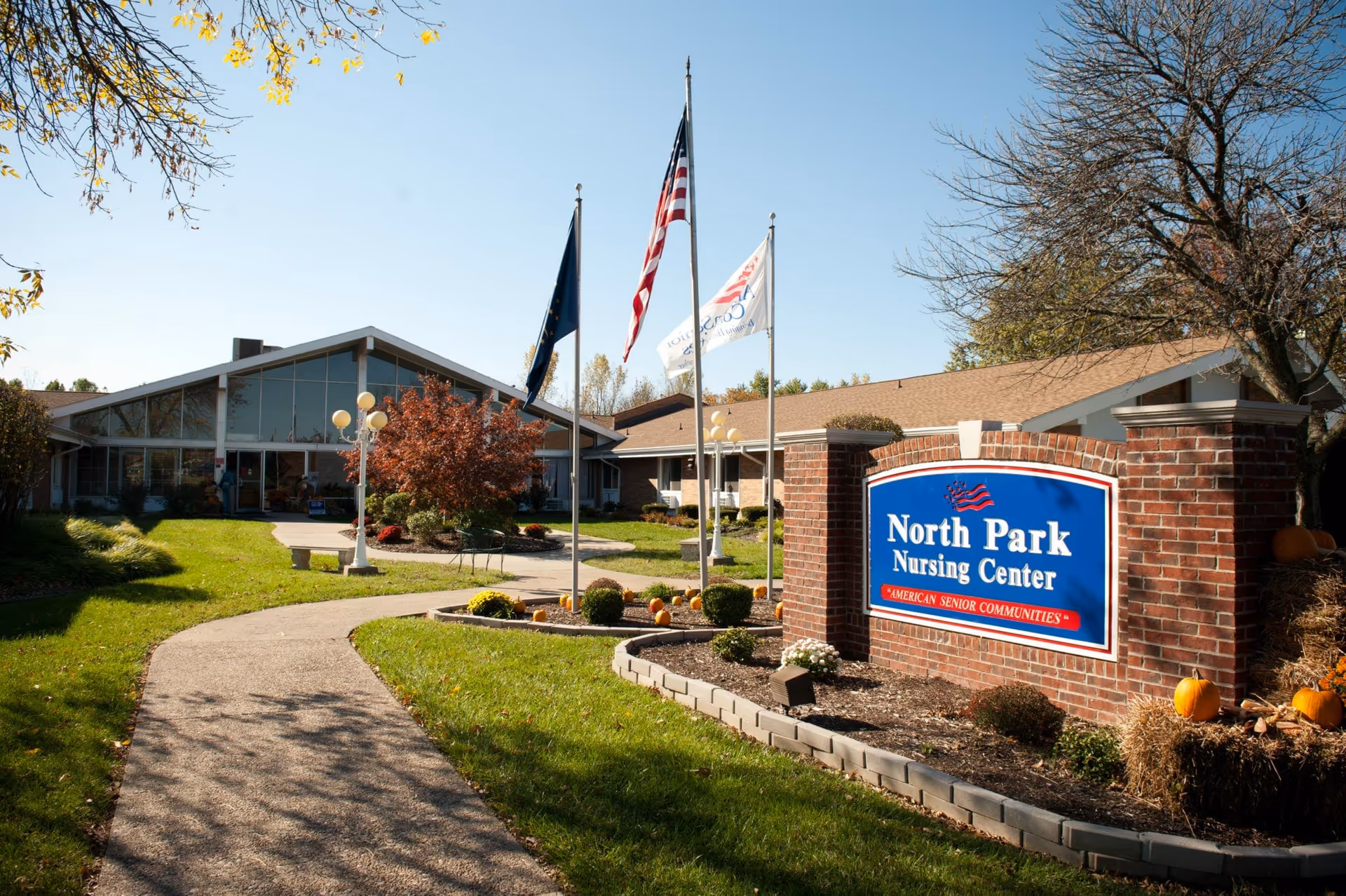 Exterior view of North Park Nursing Center with a brick sign displaying the facility's name. The building has large windows and a peaked roof. There are three flagpoles with flags in front of the building, a curved walkway leading to the entrance, green grass, trees with autumn leaves, and pumpkins decorating the landscaping.