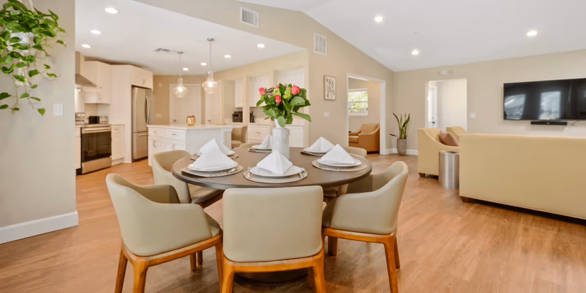 A modern open-concept interior space featuring a round dining table set with six chairs and neatly folded white napkins on plates. In the background, there is a kitchen with white cabinetry, stainless steel appliances, and pendant lights hanging over a kitchen island. To the right, a living area with beige sofas and a wall-mounted flat-screen TV is visible. The room has wooden flooring and neutral-colored walls with some plants and framed artwork.