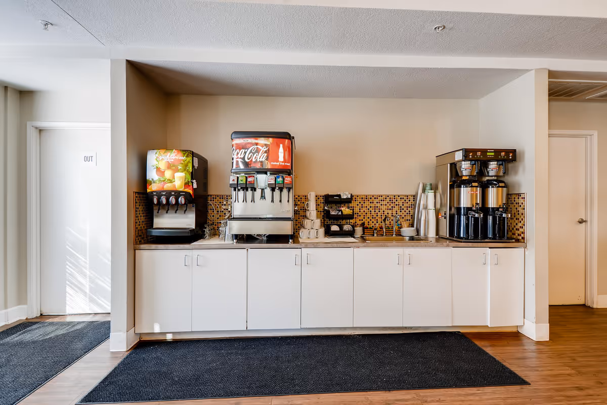 A beverage station with a soda fountain, juice dispenser, coffee machines, cups and white cabinets against a tiled backsplash in a common area.