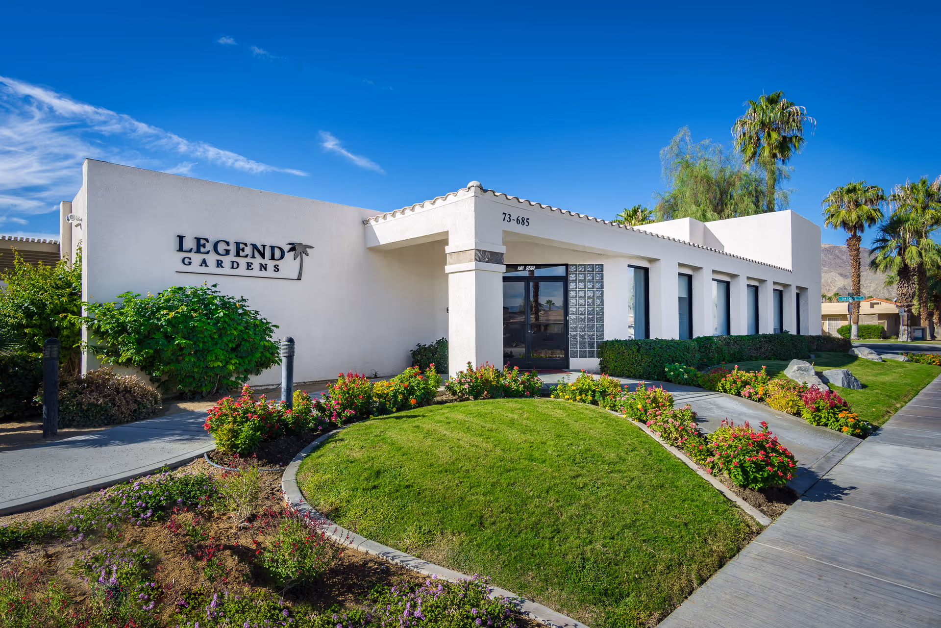 Exterior view of Legend Gardens facility showing a white building with a tiled roof, surrounded by well-maintained green lawns, colorful flower beds, palm trees, and a clear blue sky.
