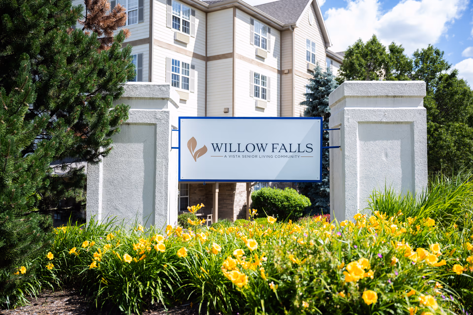Entrance sign reading "Willow Falls" in front of the senior living building surrounded by yellow flowers and greenery.