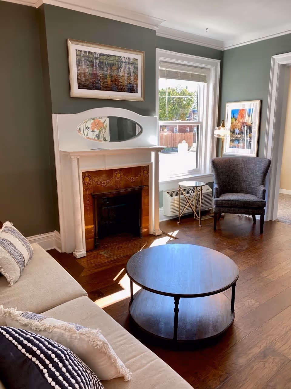 A cozy living room with a beige sofa adorned with decorative pillows, a round wooden coffee table, a dark gray armchair, and a small side table next to a window. The room features a white fireplace with a decorative tile surround and an oval mirror above it. Two framed pictures hang on the green walls, and sunlight streams through the window onto the wooden floor.