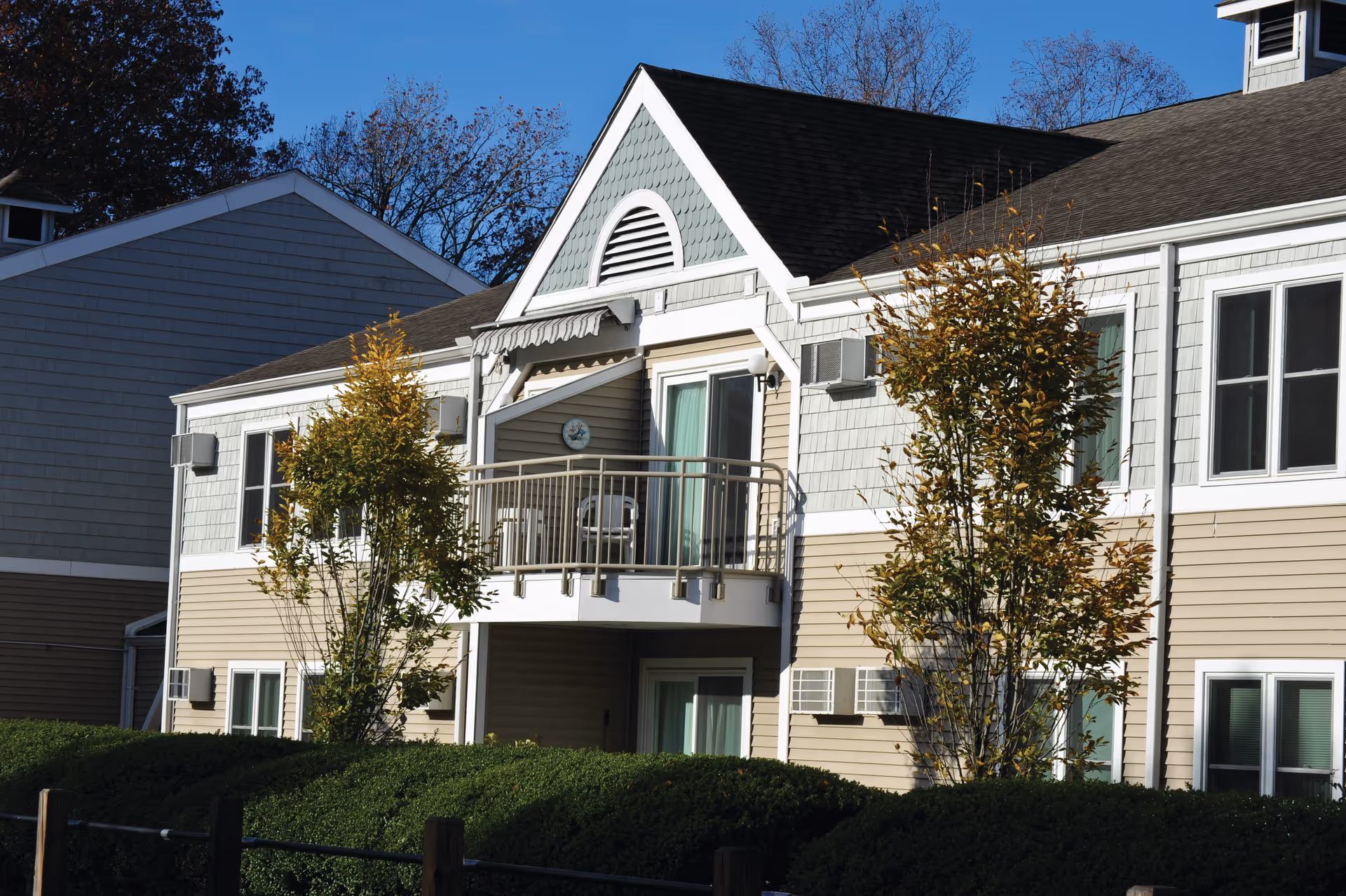 Front exterior of a two-story beige residential building with a small balcony, windows, and landscaped shrubs and trees.
