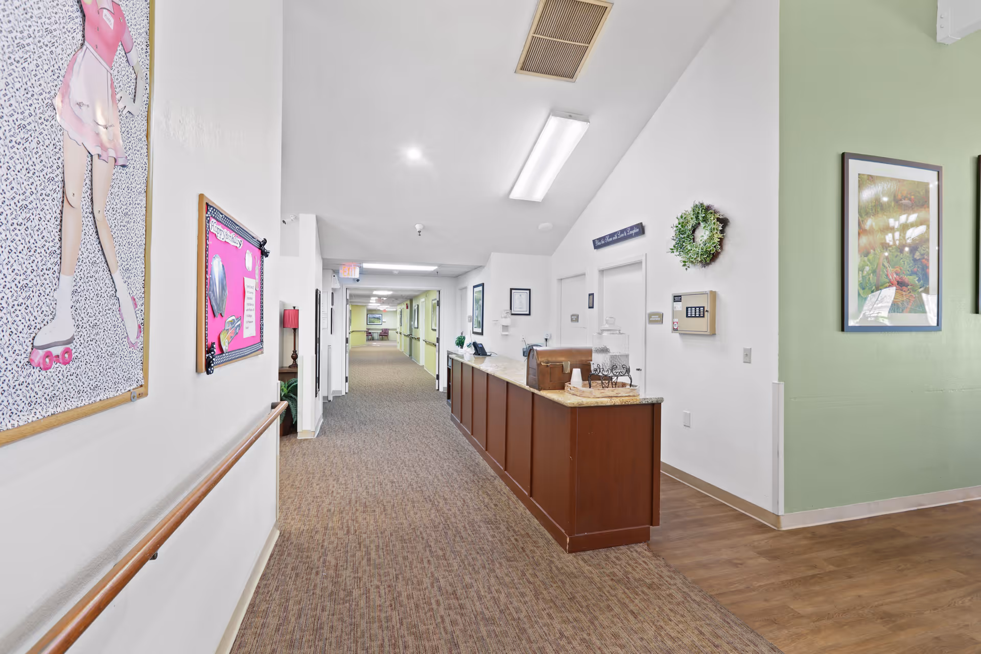 A well-lit assisted living hallway with a reception desk on the right, framed artwork and bulletin boards along the walls, and a long carpeted corridor.