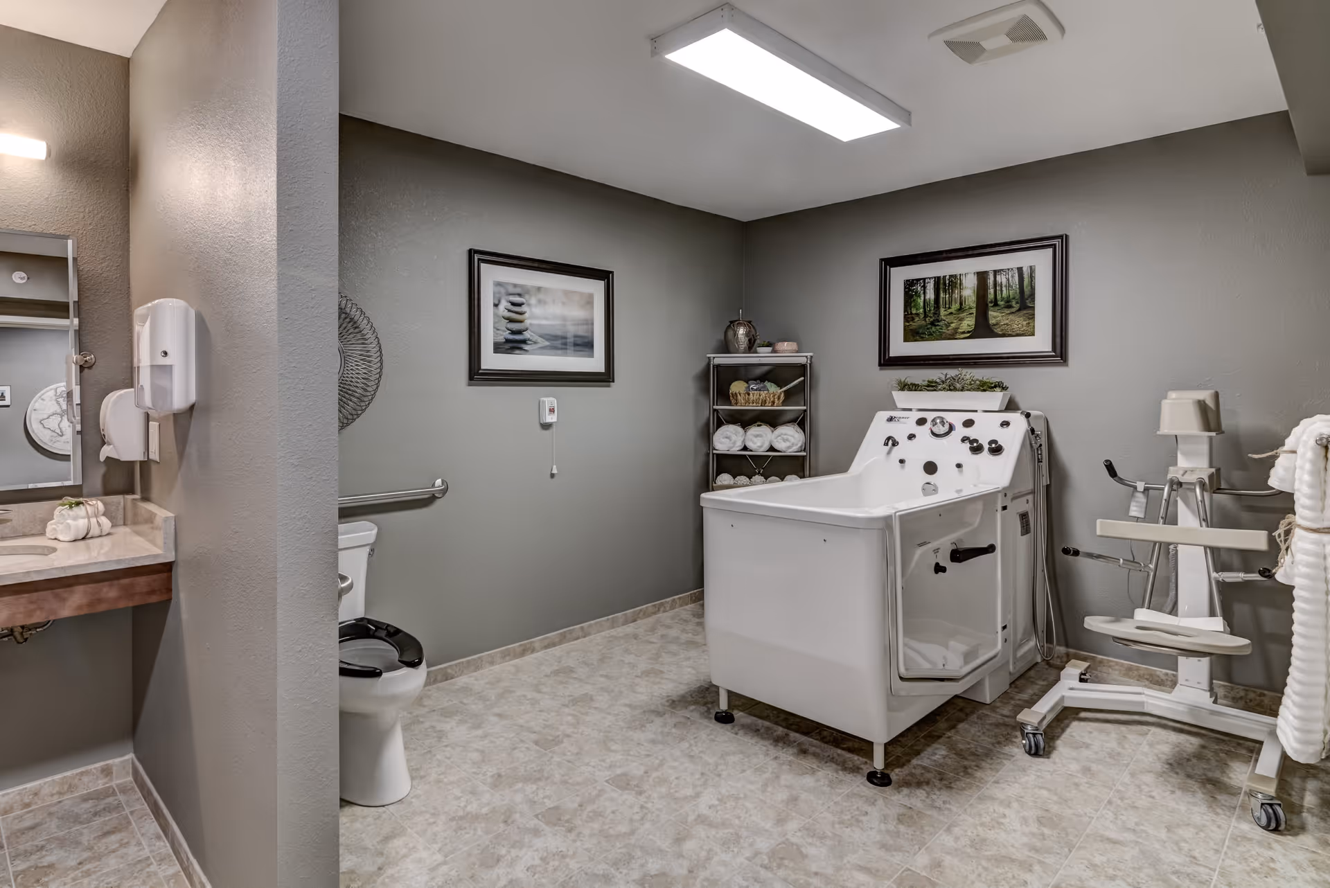A spacious bathroom with beige tiled floor and gray walls. The room features a white toilet with a black seat, a white walk-in bathtub with multiple controls, and a mechanical lift chair on wheels. There is a metal shelving unit holding rolled white towels and decorative items. Two framed pictures hang on the walls, one showing stacked stones and the other a forest scene. A sink with a countertop and mirror is partially visible on the left side.