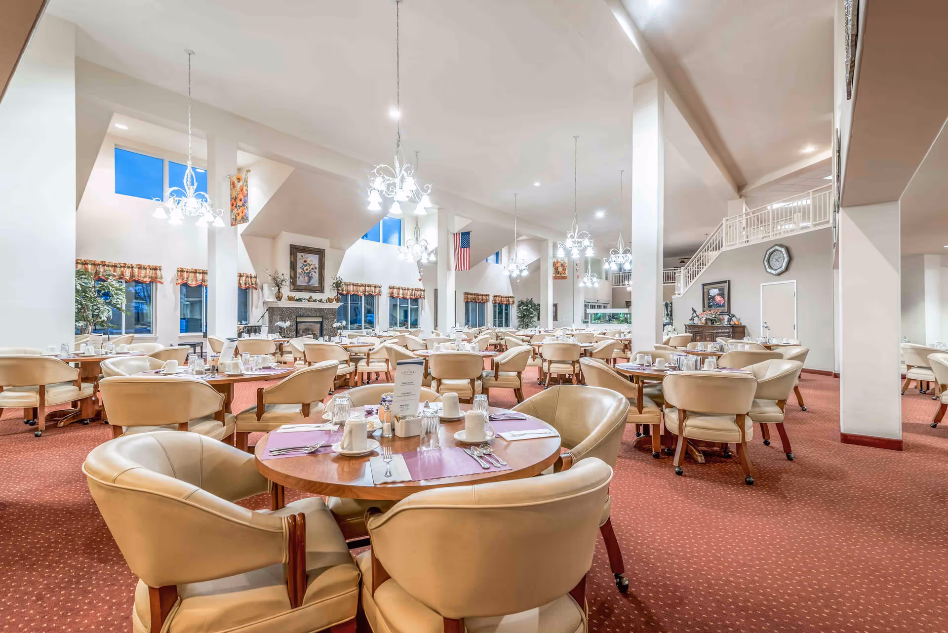 Spacious senior living dining room with round tables set for meals, beige upholstered chairs, chandeliers, a fireplace and high ceilings.