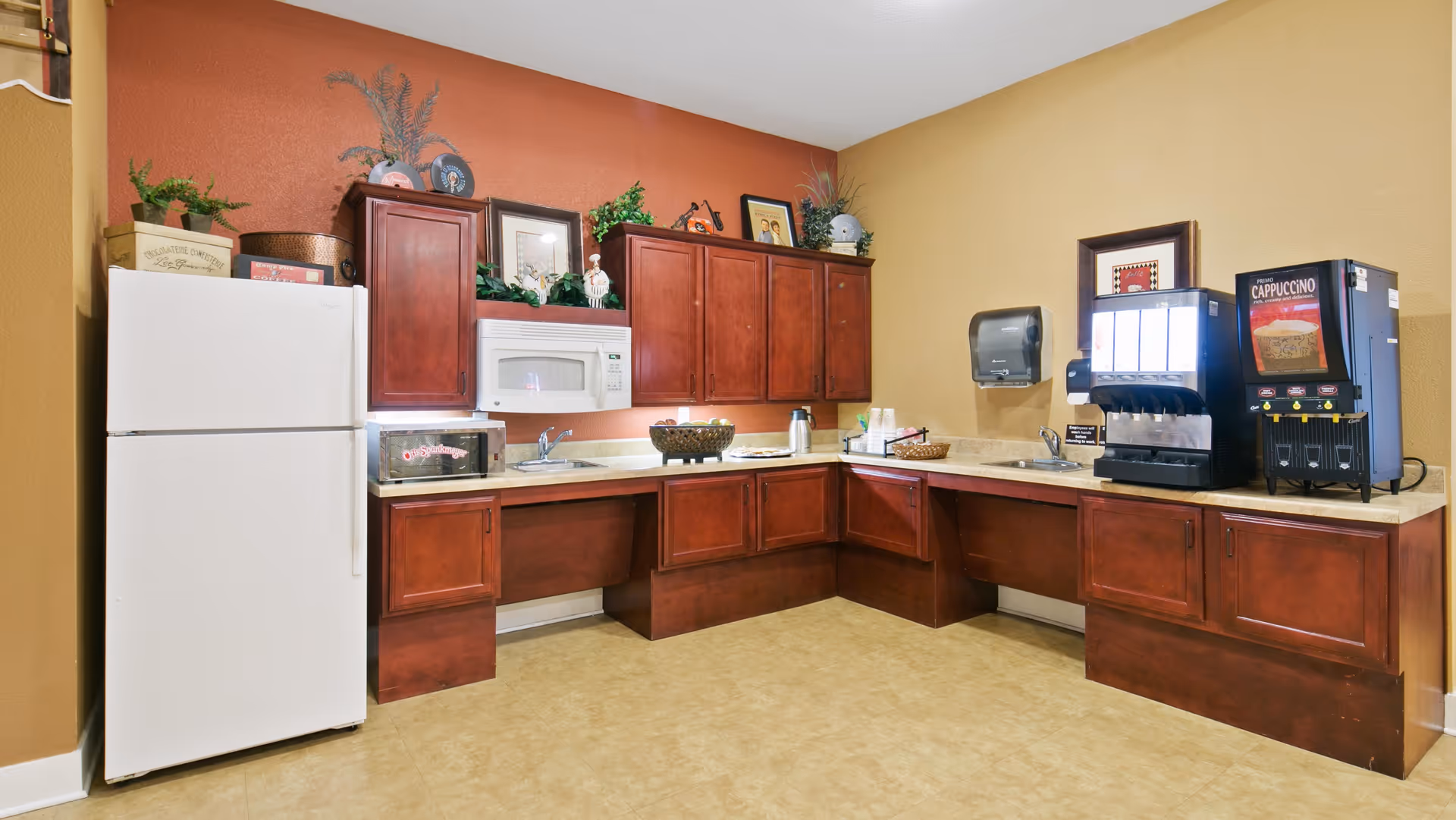 A kitchen area with wooden cabinets, a white refrigerator, a microwave, a toaster oven, two sinks, a coffee machine, and a cappuccino dispenser. The walls are painted in warm tones of orange and beige, and there are decorative plants and framed pictures on top of the cabinets.