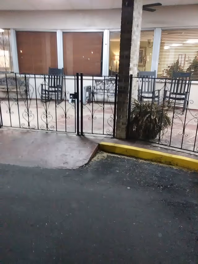 Outdoor patio area at night with metal gate and railing, two rocking chairs, a bench, potted plants, and windows with blinds showing interior lighting.