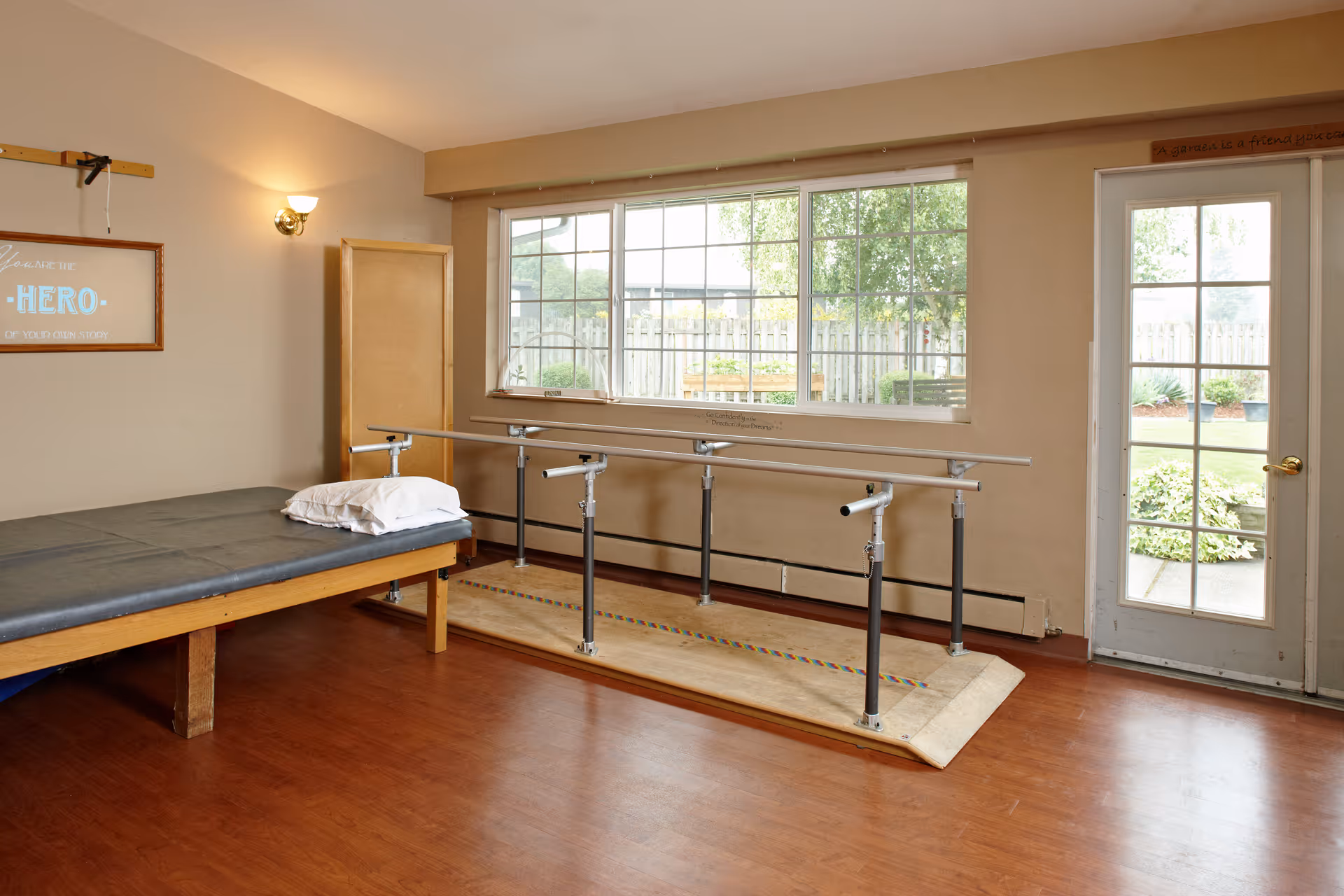A therapy room with a padded treatment table on the left, a set of parallel bars in the center for walking exercises, a large window showing a fenced garden outside, and a glass door leading to the outdoor area. The room has wooden flooring and beige walls with a framed motivational sign on the wall.