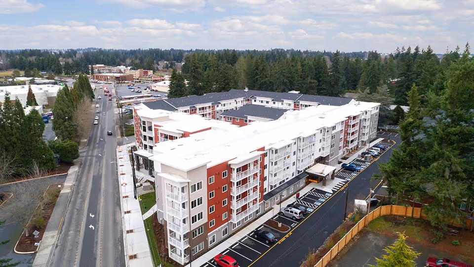 Aerial view of The Reserve At Lynnwood, a multi-story residential building surrounded by trees and parking lots, located next to a road with light traffic and commercial buildings in the background.