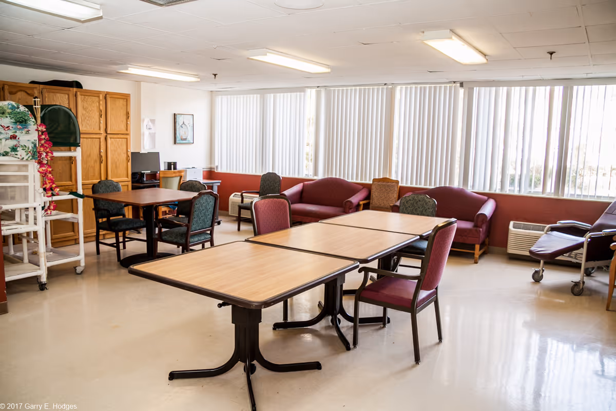 Bright communal activity/dining room with tables, chairs, sofas and large windows with vertical blinds.