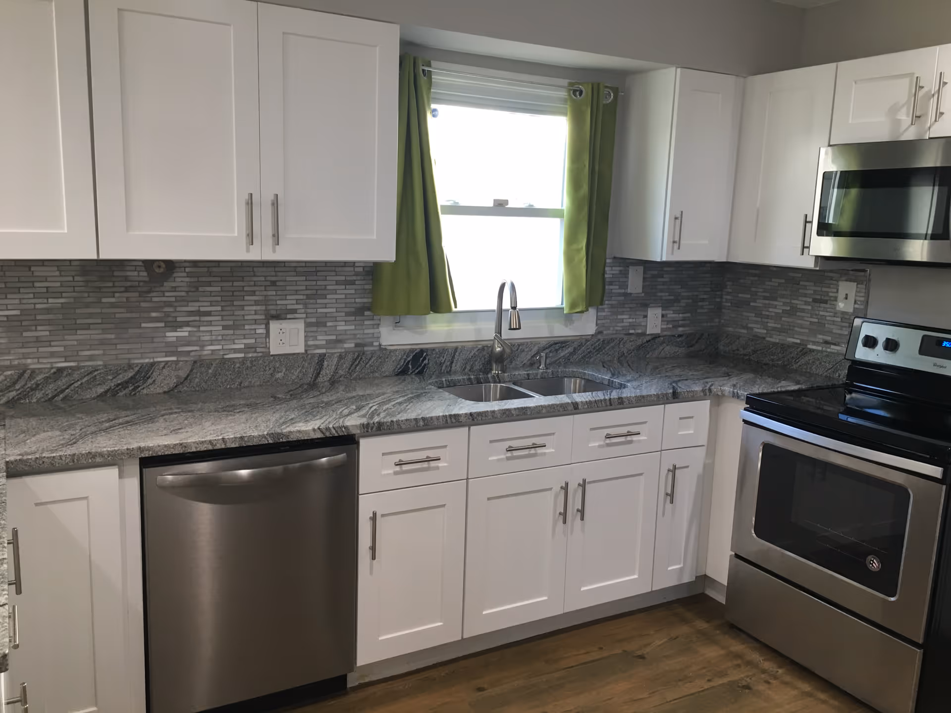 Modern kitchen with white cabinets, gray granite countertops, stainless steel appliances, and a window with green curtains above a double sink.
