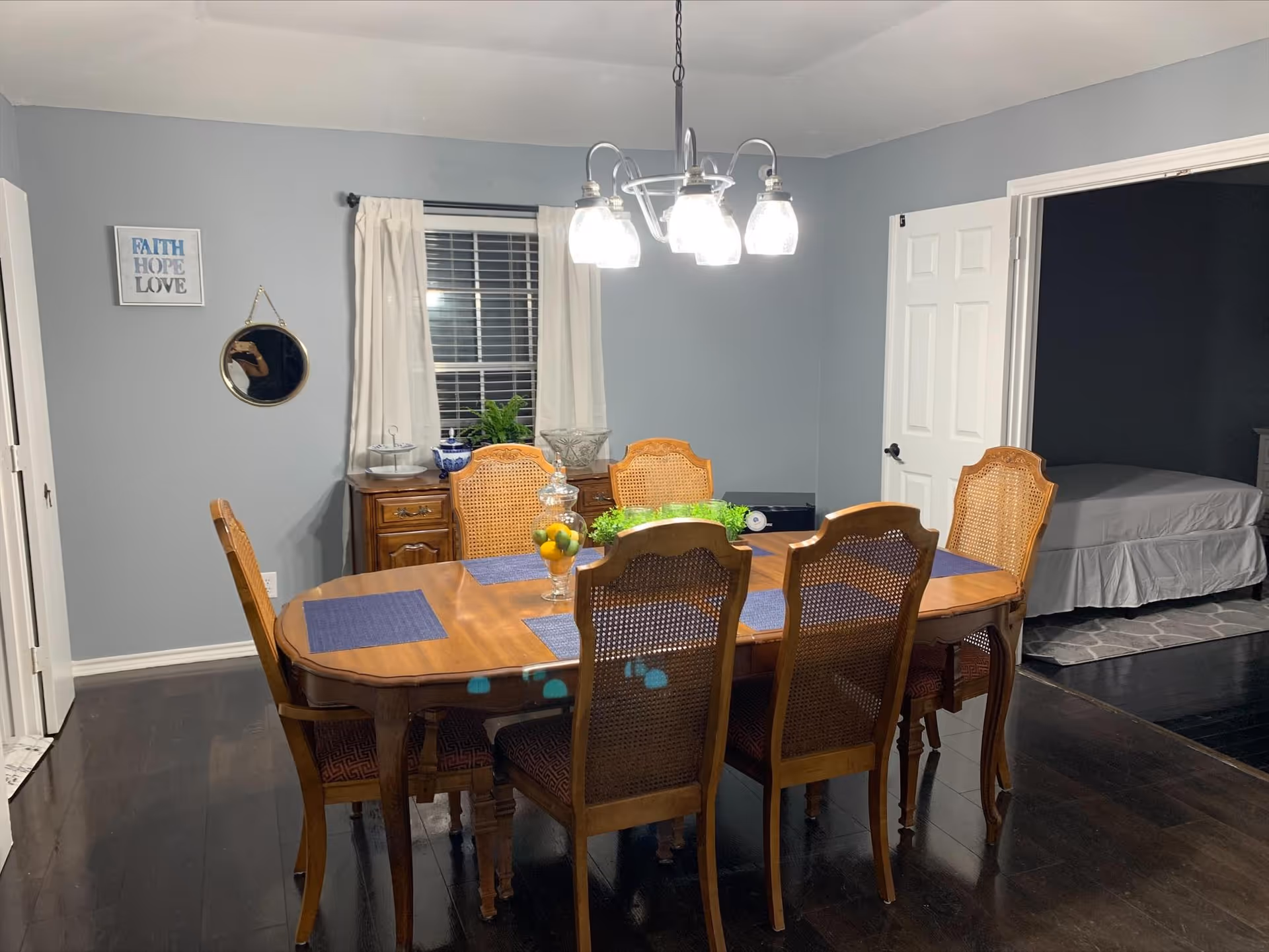 A dining room with a wooden table and six chairs with woven backs. The table has blue placemats and a centerpiece with lemons and greenery. A chandelier with five lights hangs above the table. In the background, there is a window with white curtains, a wooden sideboard with decorative items, and an open door leading to a bedroom with a bed and gray bedding. The walls are painted light gray and the floor is dark wood.