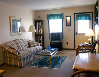 Small living room with a striped sofa, coffee table on a green rug, TV under a window, lamps, and a table near the door.