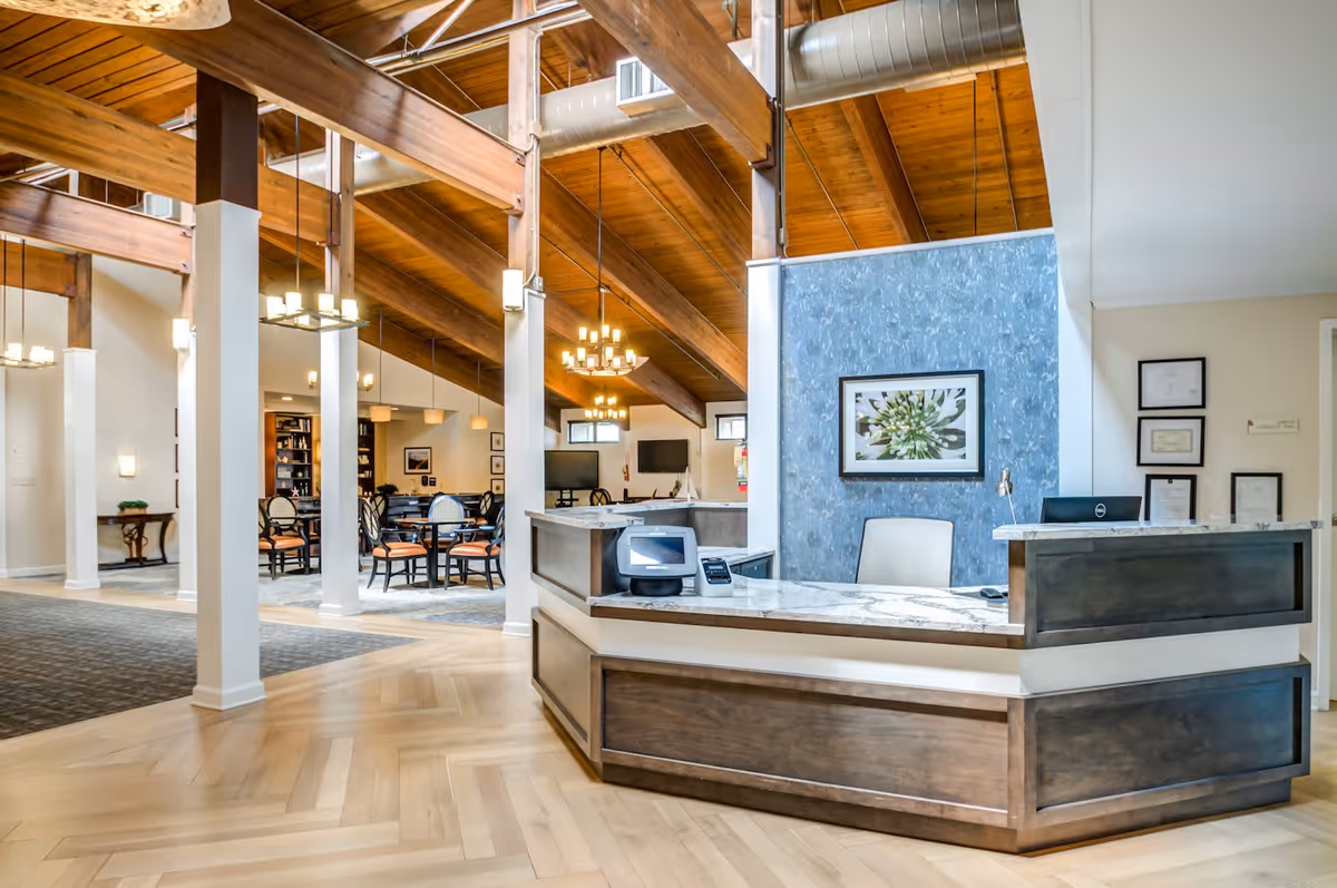 A spacious reception area in a senior living facility with a wooden ceiling featuring exposed beams and modern chandeliers. The reception desk has a marble countertop and dark wood paneling. Behind the desk is a blue textured wall with a framed floral artwork. To the left, there is a seating area with tables and chairs, and bookshelves in the background.