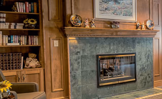 Cozy interior corner featuring a green-tiled fireplace with a wooden mantel decorated with small figurines and a framed picture above. To the left, there is a built-in wooden bookshelf filled with books and decorative items, and part of a comfortable armchair is visible.