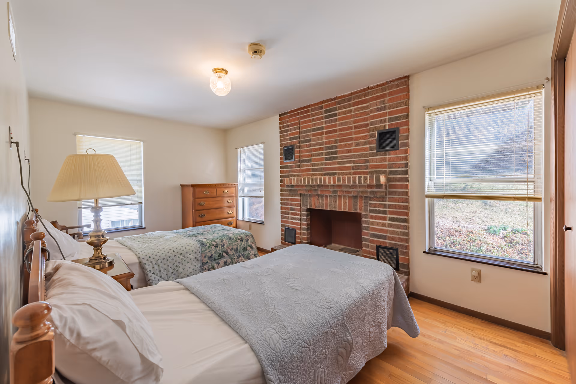 A bedroom with two single beds, each with a quilted bedspread. There is a wooden nightstand with a lamp between the beds. The room features a brick fireplace on one wall and three windows with blinds allowing natural light to enter. A wooden dresser is positioned against the far wall.