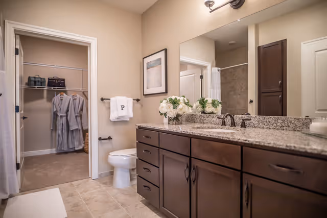 A clean and modern bathroom featuring a granite countertop with dark wood cabinets, a large mirror, a white toilet, and a towel rack with a white towel embroidered with the letter 'P'. In the background, there is an open doorway leading to a closet with hanging robes and shelves. The bathroom has beige walls and tiled flooring.