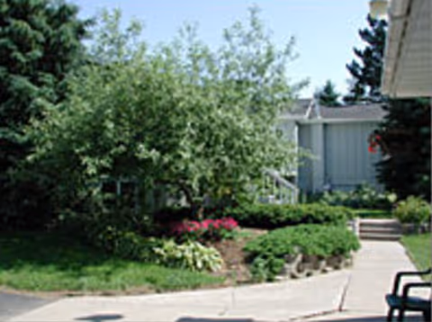 A landscaped outdoor area with a variety of green shrubs, a small tree, and flowering plants next to a paved walkway. Part of a building with light blue siding and a white roof edge is visible on the right side, along with some outdoor chairs.