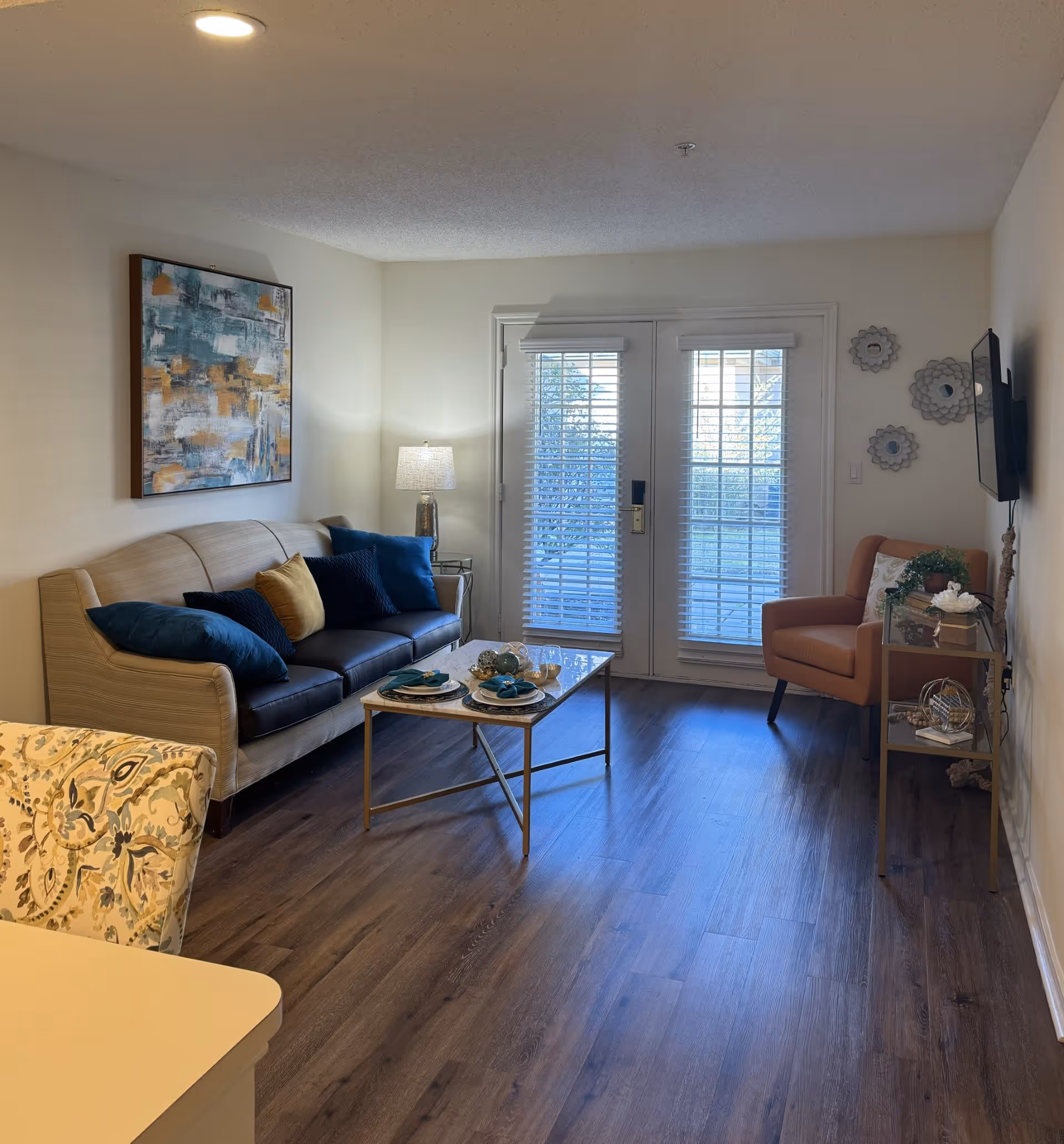 A cozy living room with a beige sofa adorned with blue and yellow cushions, a glass coffee table set with plates and napkins, a patterned armchair partially visible, a single orange armchair near a small side table with decorative items, a wall-mounted TV, and double glass doors with white blinds letting in natural light.