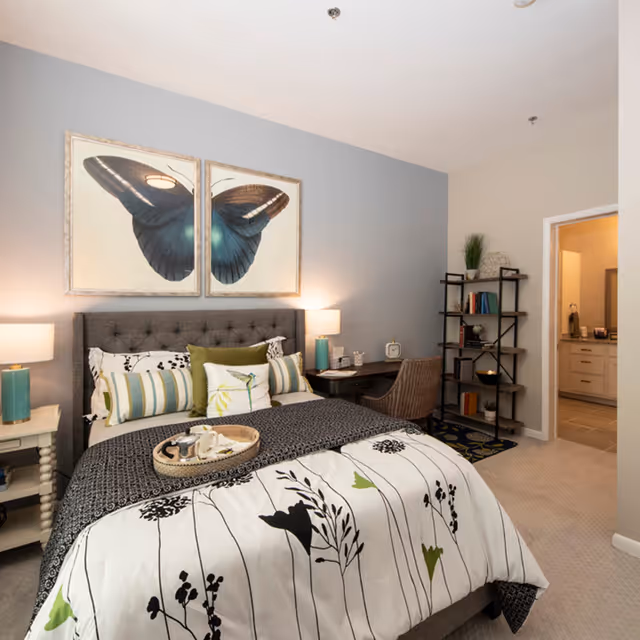 A cozy bedroom with a gray upholstered bed featuring a white and black floral patterned comforter and multiple decorative pillows. Above the bed are two framed pictures of a large blue butterfly. On the left side of the bed is a white nightstand with a turquoise lamp. On the right side, there is a small desk with a chair, a lamp, and some decorative items. A black shelving unit with books and decor is against the wall near an open doorway leading to a bathroom.