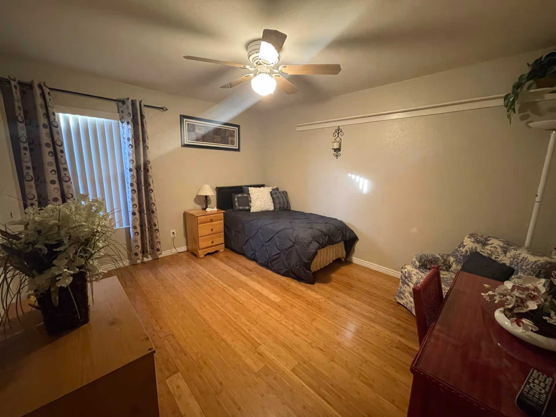 A cozy bedroom with a single bed covered in a dark comforter and multiple pillows. There is a wooden nightstand with a lamp next to the bed. A framed picture hangs on the wall above the bed. The room has wooden flooring, a ceiling fan with a light, and a window with vertical blinds and patterned curtains. A floral armchair and a red desk with a flower arrangement and a remote control are also visible.