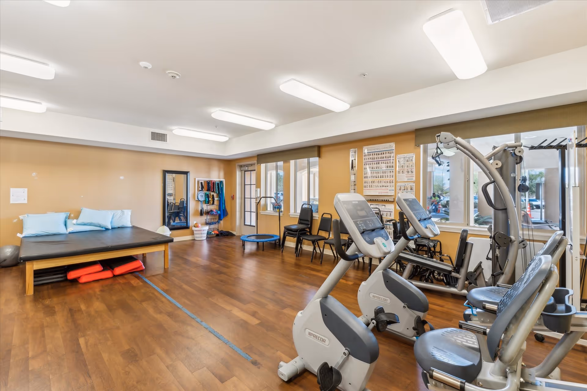 A well-lit exercise room with wooden flooring featuring stationary exercise bikes, a multi-function gym machine, a padded table with blue pillows, a small trampoline, chairs, and exercise equipment hanging on the wall. Large windows let in natural light.