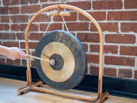 A person striking a large gong suspended on a wooden stand against a brick wall background.