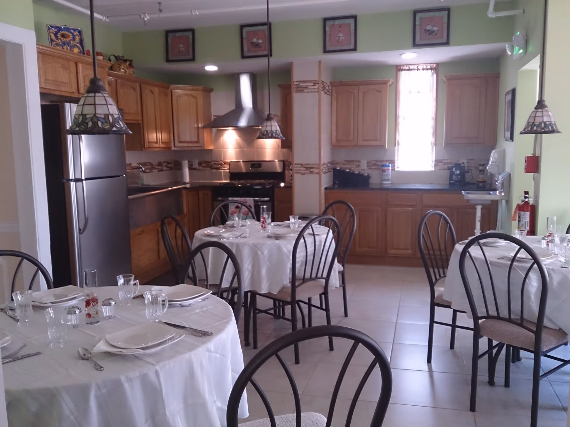 Dining area with round tables set with white tablecloths and chairs in front of a kitchen with wooden cabinets and stainless appliances.
