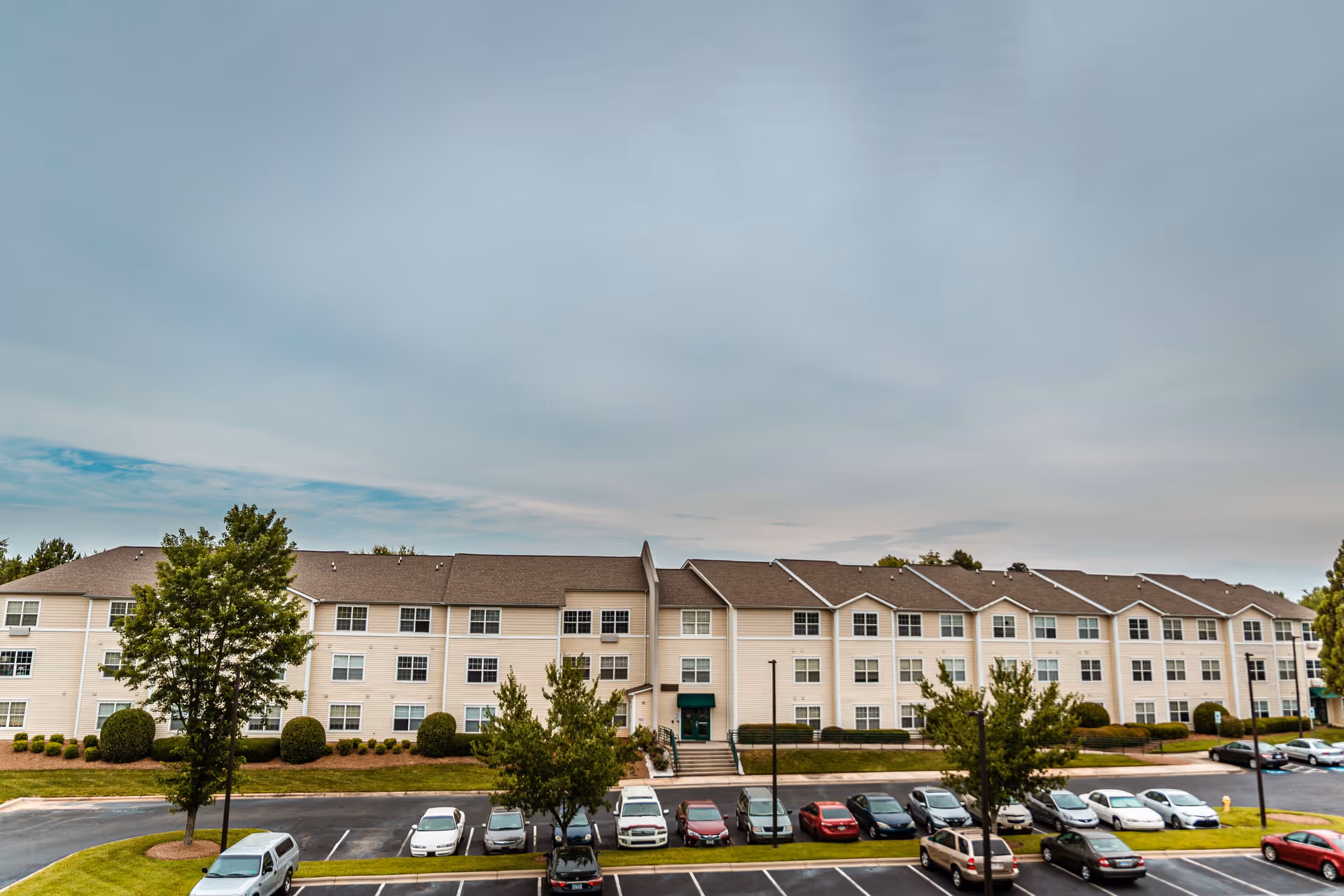 Front view of a three-story beige senior living building with landscaped grounds and a parking lot of cars under a cloudy sky.