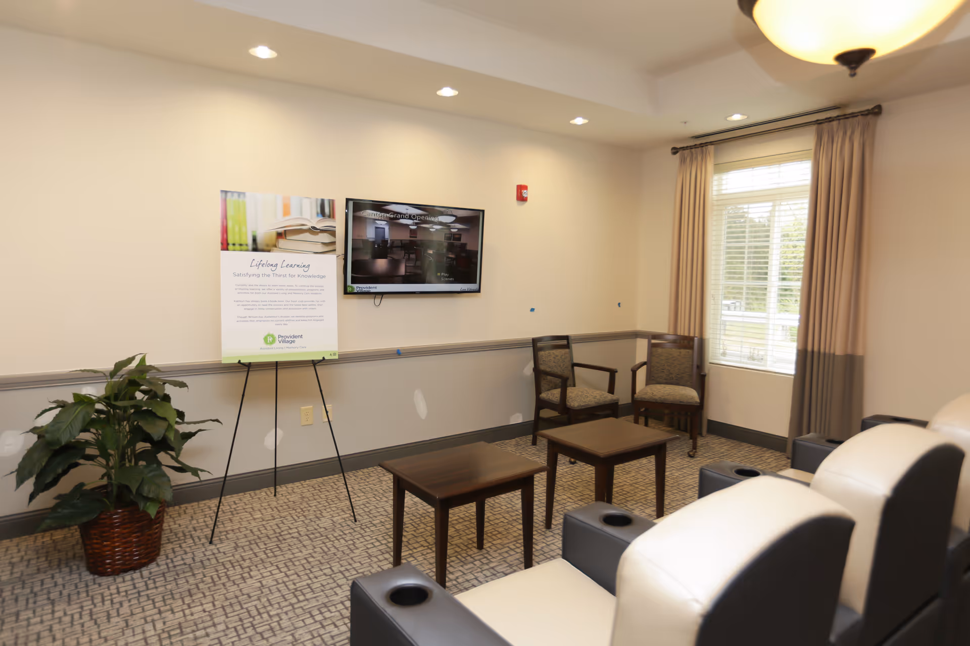 A small lounge area with beige and black cushioned chairs arranged in rows facing a wall-mounted TV. There are two wooden tables and two upholstered chairs near a window with beige curtains. A potted plant is placed on the left side, and a sign on an easel stands next to the TV. The room has recessed ceiling lights and a patterned carpet.