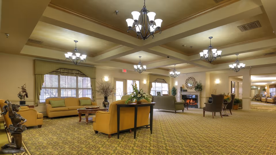 Spacious senior living community lounge with multiple seating areas including yellow and green sofas, armchairs, a fireplace with a decorative mirror above it, chandeliers hanging from a coffered ceiling, and large windows with valances letting in natural light.