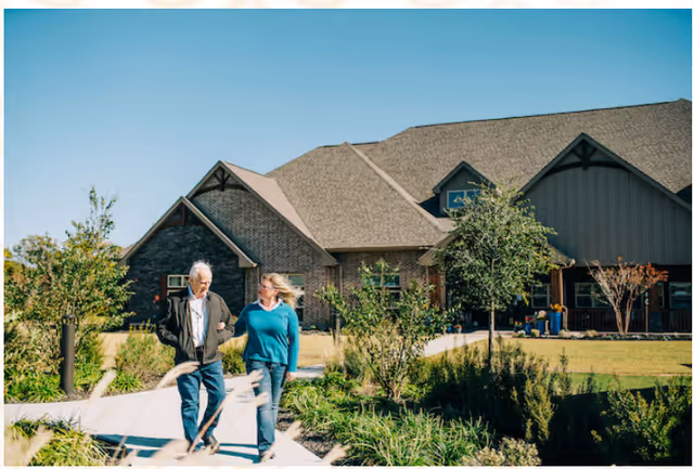 Two people walking along a landscaped path in front of a large single-story assisted living building.
