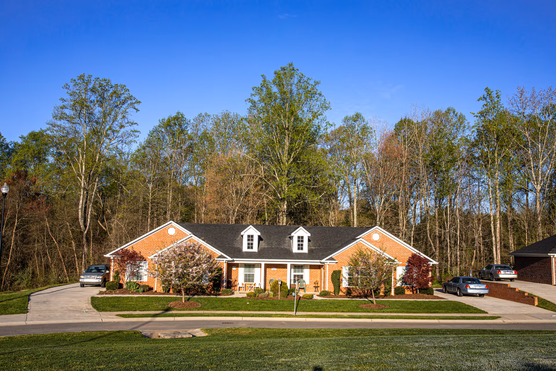 A single-story brick retirement cottage with a black roof, white window shutters, and two dormer windows. The cottage is surrounded by a well-maintained lawn, small trees, and shrubs. There are driveways on both sides with parked cars. Tall trees and a clear blue sky are visible in the background.