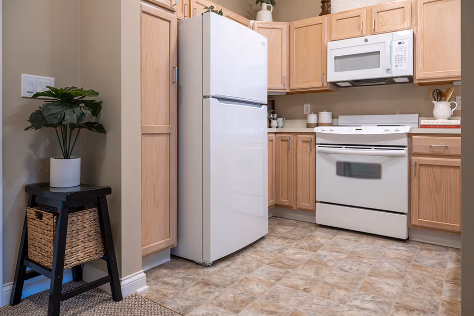 A clean kitchen featuring light wood cabinets, a white refrigerator, a white stove with oven, and a white microwave mounted above the stove. There is a small black side table with a woven basket and a potted plant on top, positioned against a beige wall. The floor has a patterned tile design.