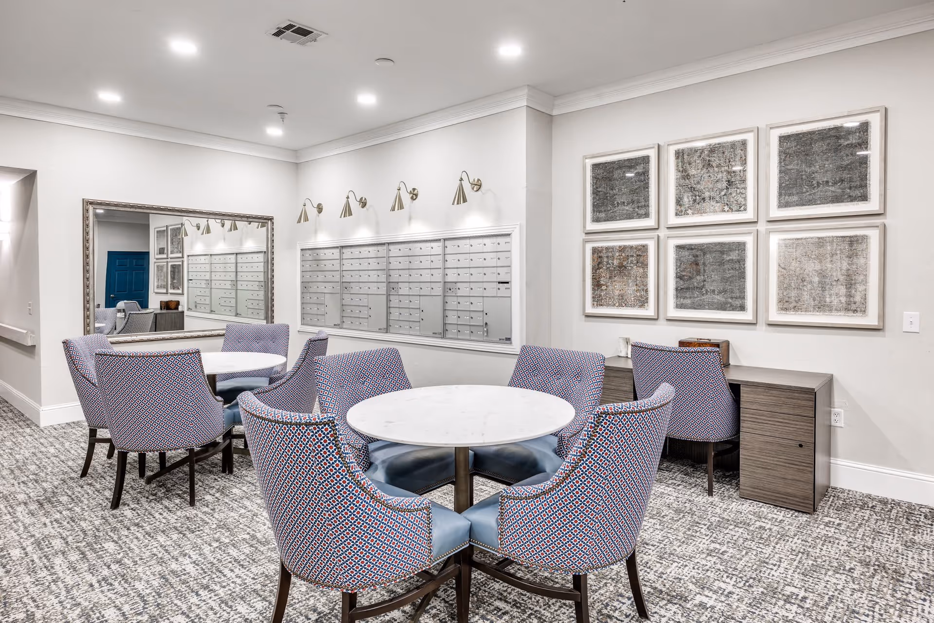 A well-lit interior room with two round white marble tables surrounded by patterned upholstered chairs. On the wall, there are multiple mailboxes with individual compartments and above them are five wall-mounted lights. A large mirror is mounted on the adjacent wall reflecting part of the room. There is also a wooden desk with two chairs and six framed abstract artworks hanging above it. The room has a carpeted floor with a gray and white pattern.