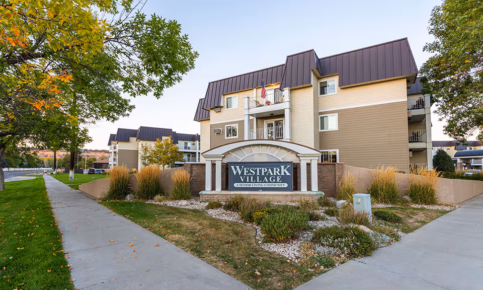 Exterior view of Westpark Village senior living community building with a sign in front surrounded by landscaping, trees with some autumn leaves, and a sidewalk leading up to the building.
