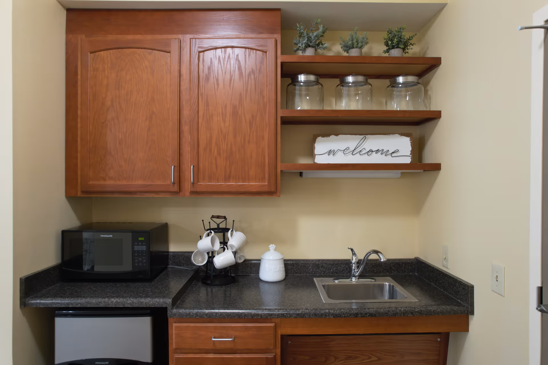 A small kitchenette area with wooden cabinets and shelves. The countertop has a microwave, a mug holder with white mugs, a white container labeled 'treats', and a stainless steel sink with a faucet. On the shelves above, there are three glass jars and three small potted plants, along with a decorative sign that says 'welcome'.