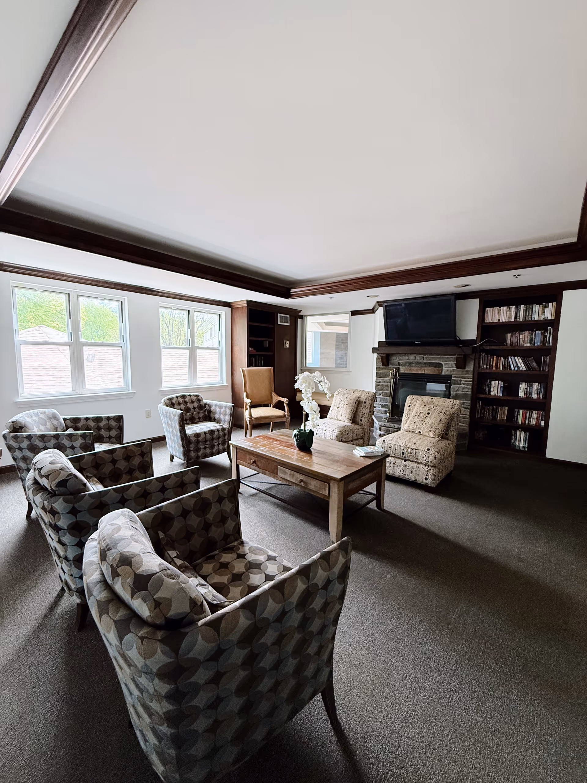 A senior living common room with patterned armchairs arranged around a wooden coffee table, a fireplace with TV, bookshelves, and windows.