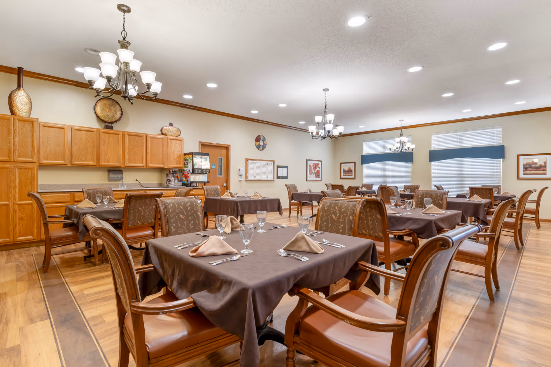 A dining room in a senior living facility with multiple tables covered in brown tablecloths, each set with beige napkins, glassware, and silverware. The room features wooden chairs with cushioned seats and backs, hardwood flooring, and overhead chandeliers. There are large windows with blinds and valances, wooden cabinets along one wall, and decorative plates and framed artwork on the walls.