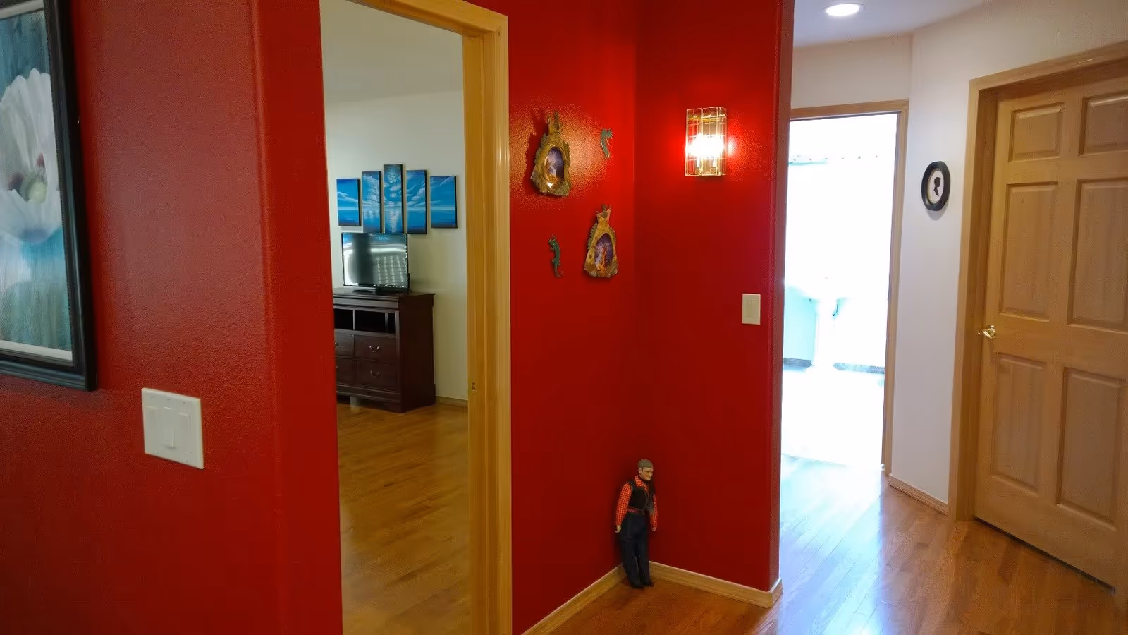 Interior view of a hallway with red and beige walls, wooden floor, and wooden doors. A small figurine stands in the corner against the red wall, which is decorated with wall art and a light fixture. Through an open doorway, a room with a TV on a wooden stand and blue wall art is visible.