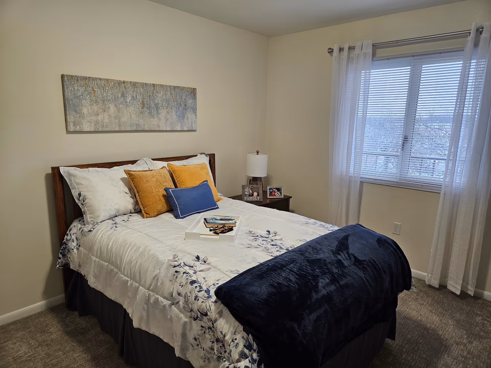 A neatly made bed in a bedroom with white and floral bedding, two mustard yellow pillows, and a blue pillow. A dark blue throw blanket is folded at the foot of the bed. There is a wooden nightstand with a lamp and framed photos next to the bed. A window with white sheer curtains lets in natural light. A textured abstract painting hangs on the wall above the bed.