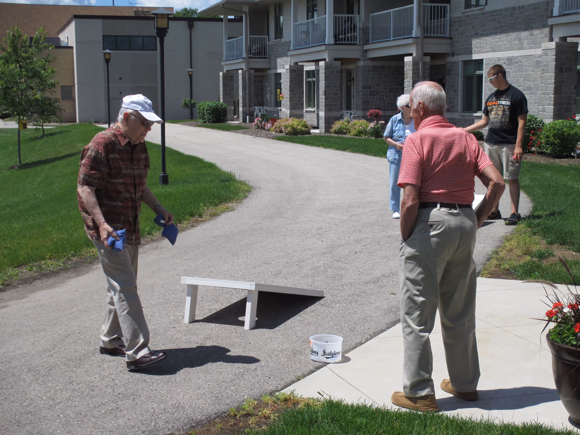 Four people outdoors at a senior living facility playing a game of cornhole on a paved path near a building with balconies and landscaped greenery.